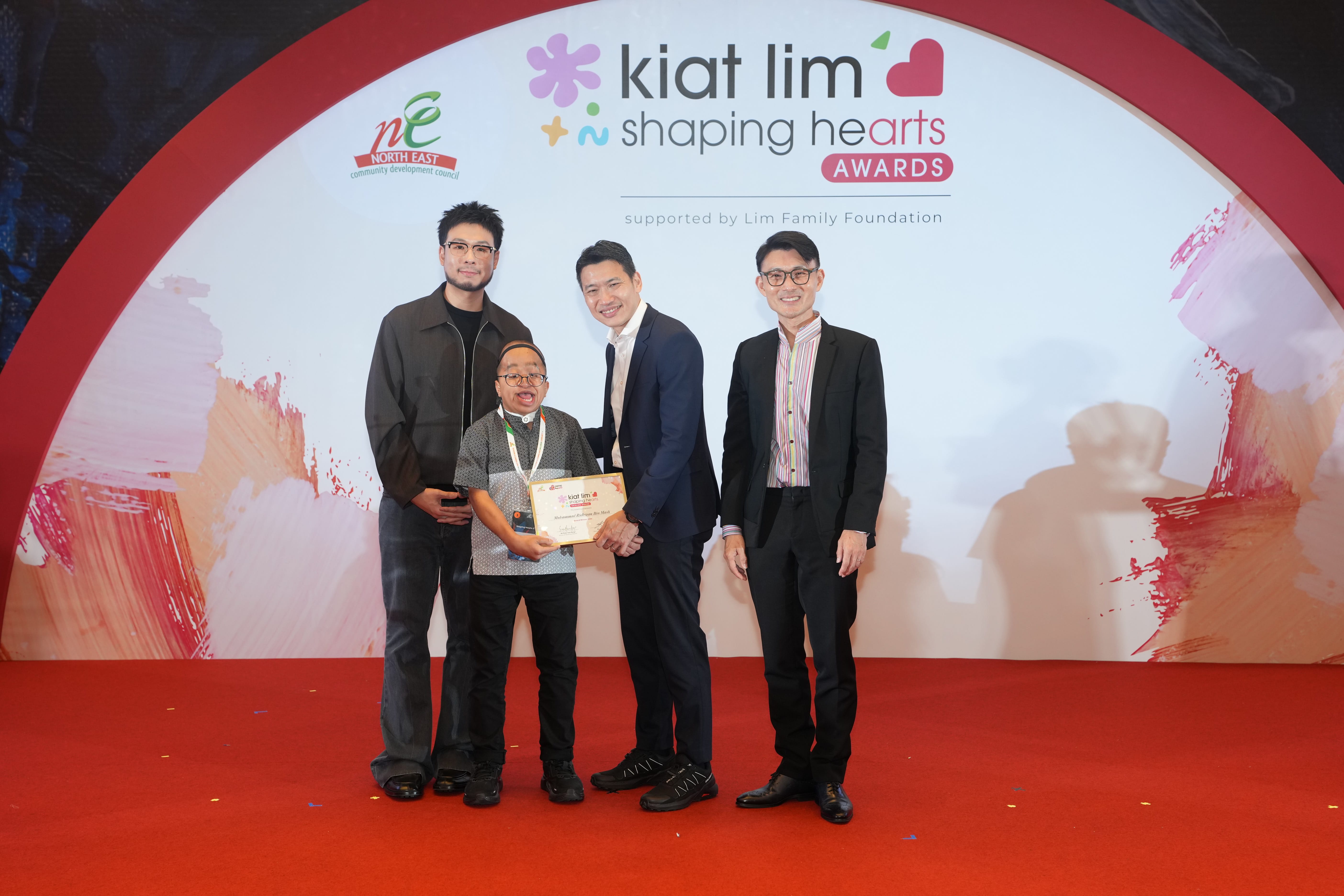 Award recipient standing on stage with the presenter, holding their certificate and posing for a group photo during the award ceremony, with a backdrop reading ‘Kiat Lim-Shaping Hearts Awards'.